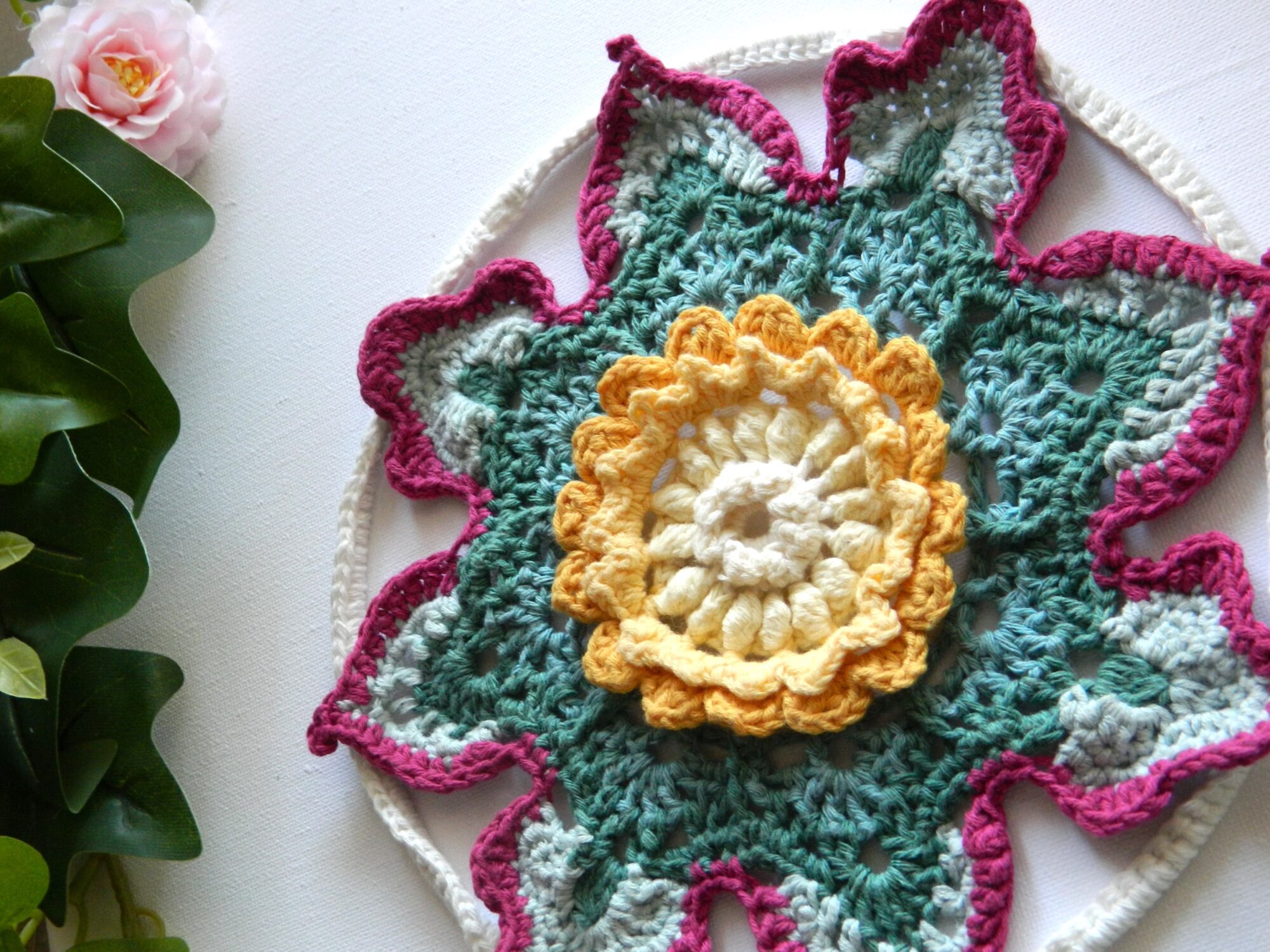 Attrape-rêves fleur au crochet BLOM - vue artistique gauche Attrape-rêves au crochet en forme de fleur dans des tons vert, crème, jaune et fuchsia.