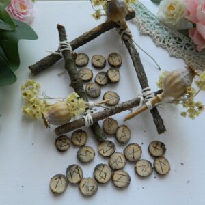 Jeu de runes en chêne artisanal – Futhark ancien Jeu de 24 runes en chêne gravées à la main, inspirées du Futhark ancien, présenté dans une mise en scène naturelle avec branches, fleurs séchées et éléments végétaux.